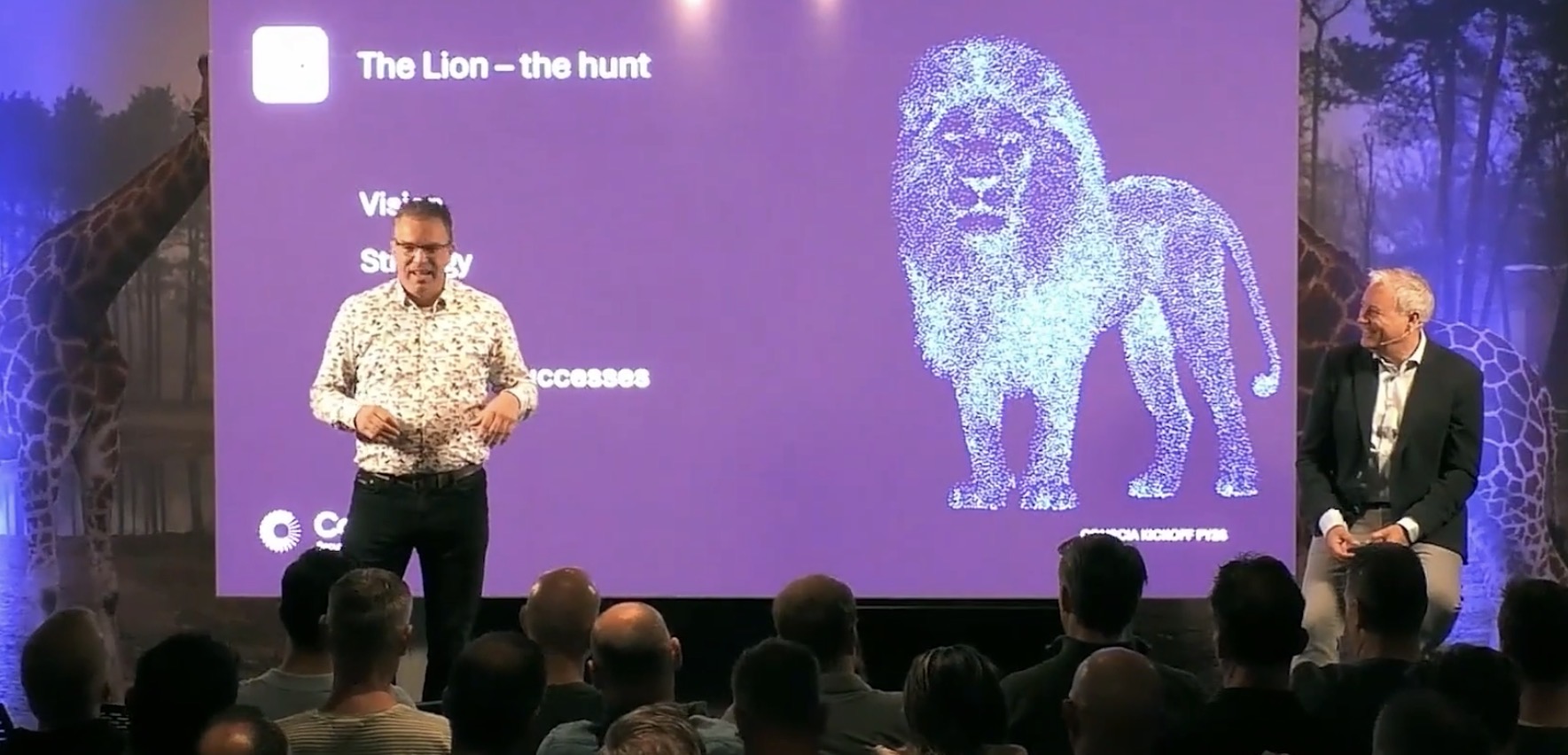 Robert van Schie spreekt op een kick-off, met een leeuw op het scherm